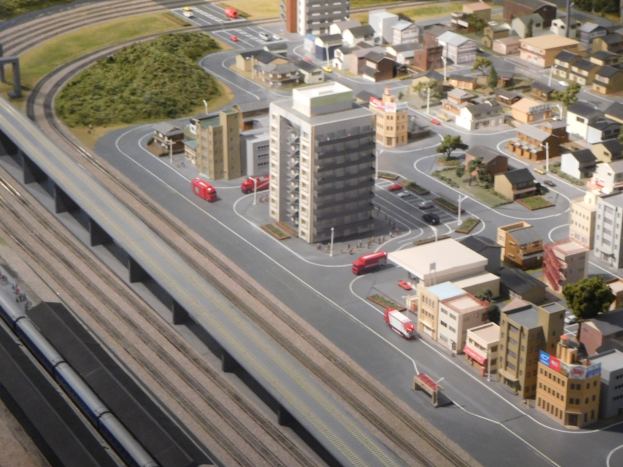 Ein Miniaturstadtmodell mit einer Bahn auf Schienen, umgeben von Gebäuden, Bäumen, Straßen, Fahrzeugen, Strommasten und Gras, mit einem Wolkenkratzer im Hintergrund.