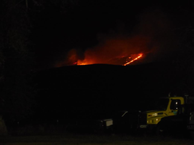 Ein Fahrzeug wird nachts mit einem Feuer im Hintergrund gezeigt.