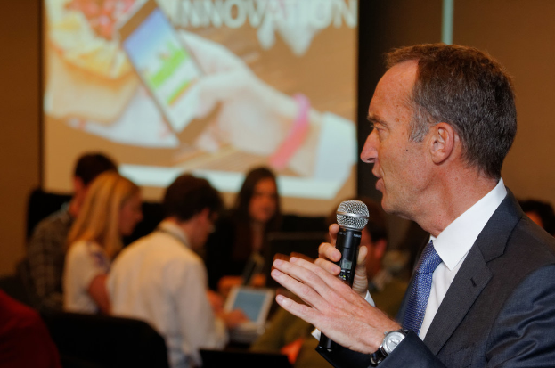 Eine Person in einem Anzug spricht in ein Mikrofon, während einige Personen hinter ihr sitzen.