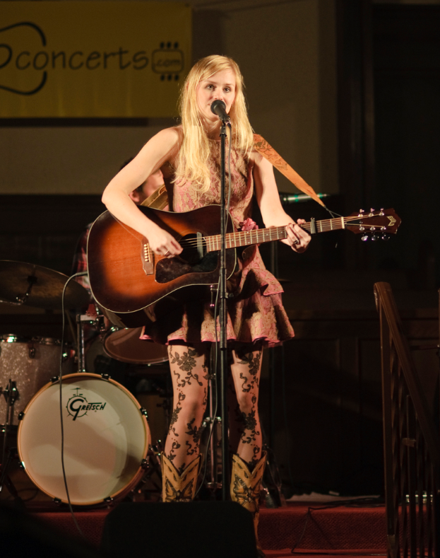 Eine Frau spielt Gitarre und singt auf der BÃ¼hne bei einem Konzert, mit einer Band und einem Bildschirm im Hintergrund.