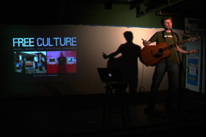 Eine Person, die eine Gitarre hält und auf einem Tisch ein Laptop sowie im Hintergrund ein Projektionsbild mit der Aufschrift "Free Culture" zu sehen ist.
