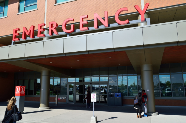 Gruppe von Menschen, die auf ein Notaufnahmelokal mit Glaswänden, Säulen und einer Namensplakette zusteuern, begleitet von Hinweistafeln und einem Mülleimer.