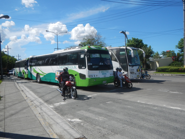 Ein grüner und weißer Shuttlebus steht am Straßenrand mit Motorradfahrern davor, ein grasbewachsener Fußweg links daneben und Gebäude, Bäume, Laternenmasten und einen klaren blauen Himmel im Hintergrund.