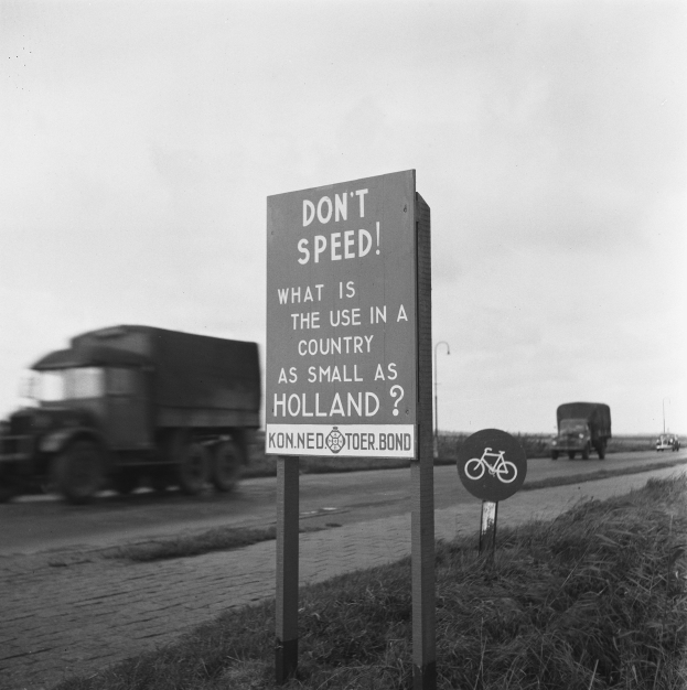 Schwarz-weißes Foto eines Lkws auf einer Straße mit einem "Fahr nicht zu schnell"-Schild im Vordergrund und anderen Fahrzeugen, Gras, Pfählen und einem klaren Himmel im Hintergrund.