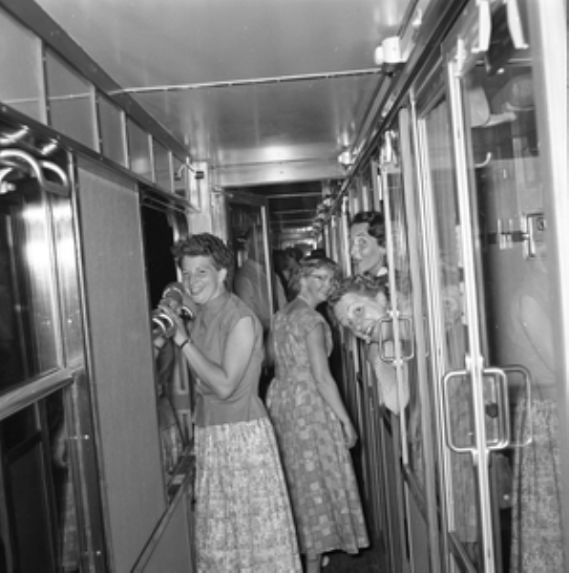 Eine Gruppe von Frauen, die auf einem schwarzen und weißen Zug mit Türen auf beiden Seiten und Lichtern öber ihnen steht.