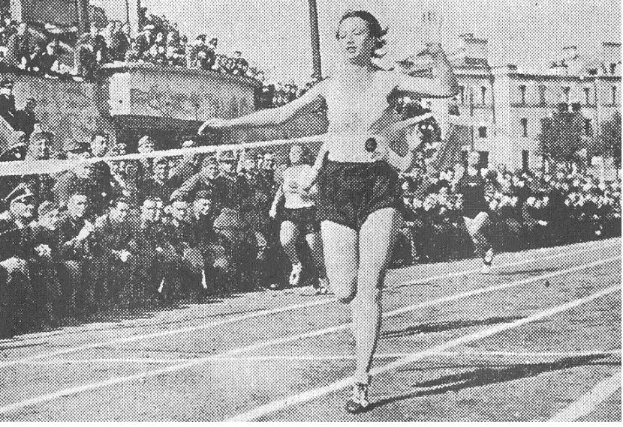 Ein Schwarz-Weiß-Foto einer Frau, die auf einer Laufbahn während des Frauen-100m-Finals bei den Olympischen Spielen 1956 läuft, mit einer Zuschauermenge und Gebäuden im Hintergrund.