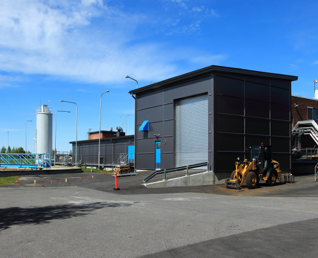 Ein großes Gebäude an einem neuen Industriepark mit einem Bagger davor, umgeben von Straßenmasten, Straßenlaternen, Kraftfahrzeugen, Absperrpollern, Geländern, Bäumen und einem bewölkten Himmel.