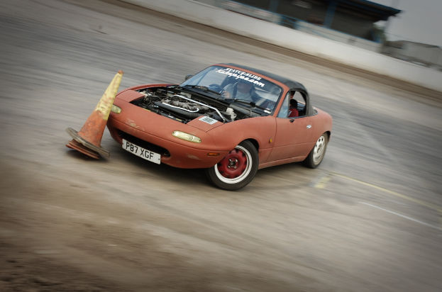 Eine Person fährt ein rotes Auto mit hoher Geschwindigkeit auf einer Rennstrecke, vor sich einen Verkehrskegel, der Hintergrund ist unscharf.