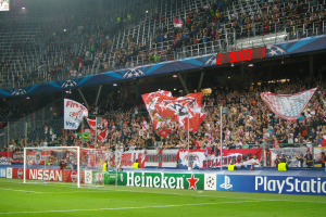 Ein Fußballspiel wird in einem hell erleuchteten Stadion mit einer großen Zuschauermenge, grünem Rasen, einem Torpfosten mit Bannern und einem Bildschirm in der oberen linken Ecke gespielt.
