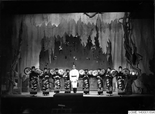 Schwarzes und weißes Bild von Darstellern auf einer Bühne in traditionellen japanischen Kimonos, die Gegenstände halten, mit einer bemalten Kulisse und 'Kimono-Show in Tokyo, Japan' Text unten.