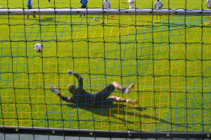 Ein Fussballer hält den Ball vor dem Tor, mit einem Netz im Vordergrund und einigen Menschen im Hintergrund sowie mehreren Bällen auf dem Boden verstreut.