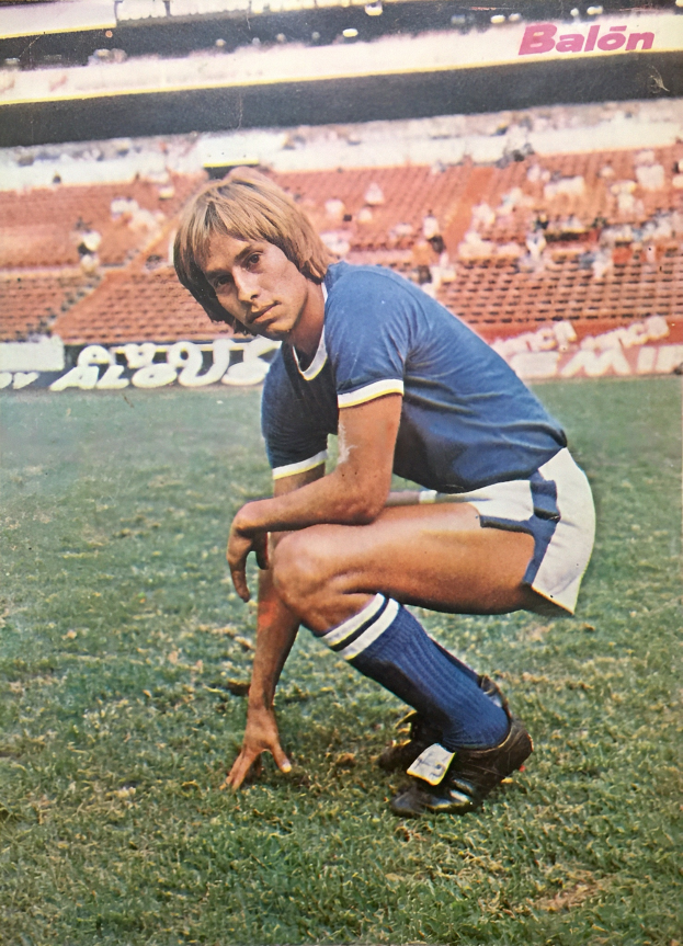 Ein Mann kniet auf einem Fußballfeld, umgeben von sitzenden Personen und Stühlen mit Text darauf, trägt Schuhe.