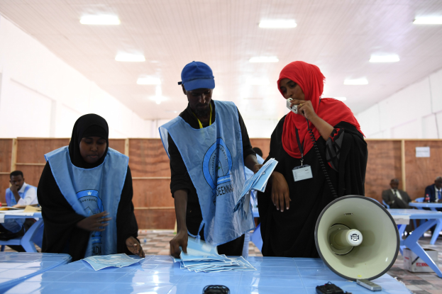 Eine Gruppe von Menschen versammelt sich um einen Tisch mit einem Megafon, Papieren und anderen Gegenständen, die scheinbar eine Wahllokalität sind, mit anderen im Hintergrund sitzend und stehend unter Deckenlampen.