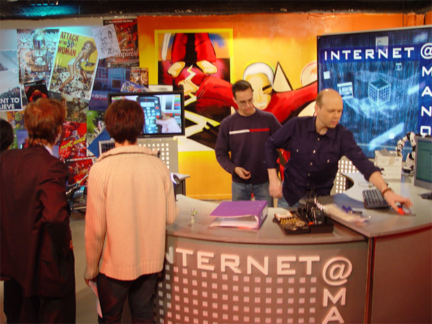 Mehrere Personen in einem Raum mit einem Tisch, der eine Akte, eine Tastatur, einen Monitor und eine Maus enthält, mit einer Wand im Hintergrund, die Plakate und eine Cartoon-Figur zeigt.