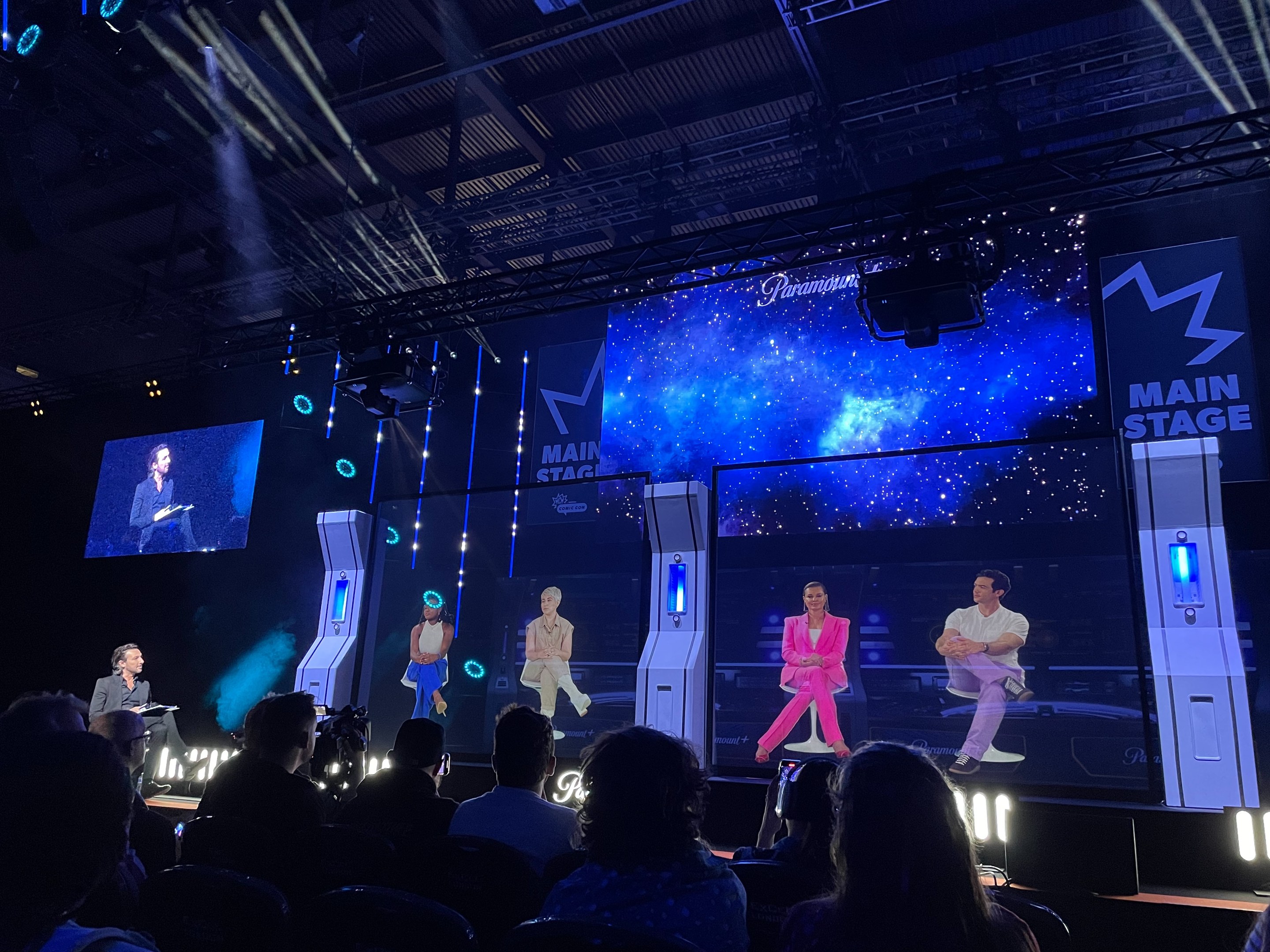 Eine Gruppe von Menschen auf einer hell erleuchteten Bühne mit Bildschirmen, die einen Redner mit Mikrofon und ein Publikum davor zeigen, über einer Decke mit Beleuchtungsequipment.