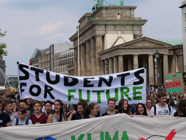 Eine Gruppe von Schülern marschiert in Berlin, die eine bunt bemalte Fahne mit der Aufschrift "Students for Future" trägt, vor dem Hintergrund von Gebäuden, Bäumen und Himmel.