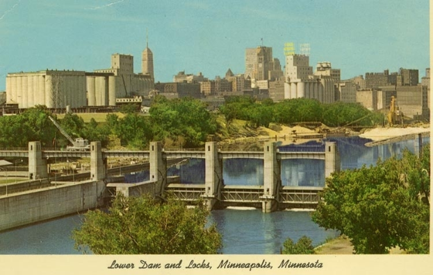 Eine Brücke spannt einen Fluss mit Bäumen und Gebäuden im Hintergrund, mit dem Text "Lower Dam and Locks, Minneapolis, Minnesota" unten.