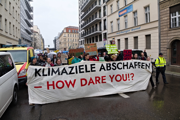 Gruppe von Menschen mit einem Banner "Klimaziele Abschaffen - Wie Wagst Du Es?" auf der Straße mit Fahrzeugen, Gebäuden, Schildern und einem klaren blauen Himmel im Hintergrund.