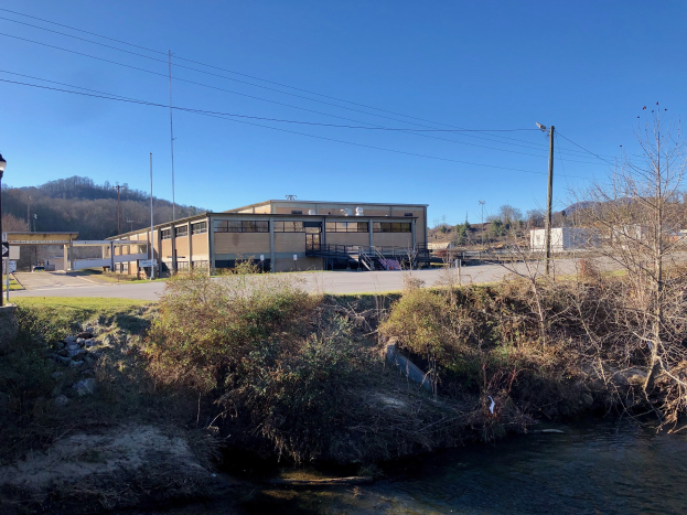 Ein Fluss fließt durch ein grünes Feld neben einem Gebäude, umgeben von Bäumen, Pflanzen, Gras, Steinen, Straßenmasten, Straßenlaternen, Strommasten, Stromkabeln, Hügeln und einem klaren blauen Himmel, wobei das Gebäude als Standort einer geplanten Wasseraufbereitungsanlage vorgeschlagen wird.