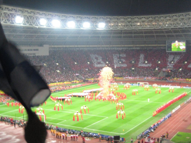Ein Stadion mit Flutlicht, Eisengittern, Zuschauern, stehenden Personen, Werbetafeln, Anzeigetafeln, Überwachungskameras, Fahnen und Deko.
