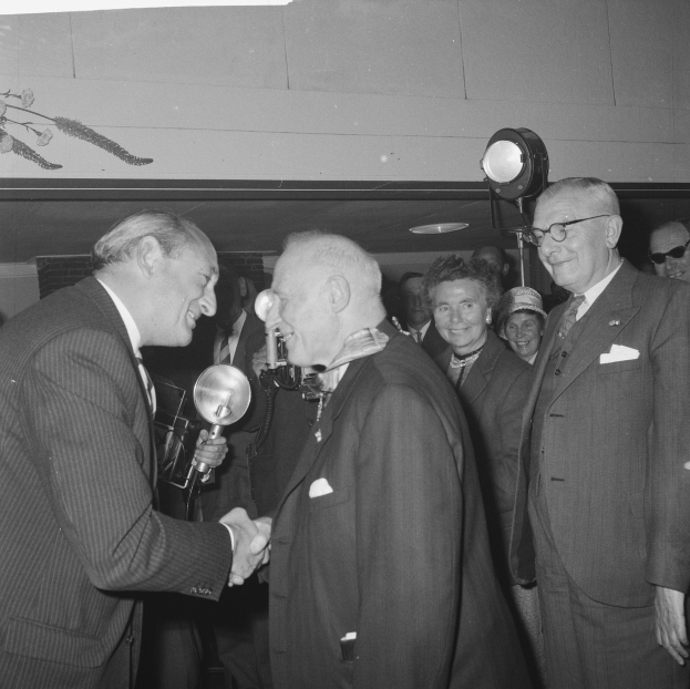 Zwei Männer schütteln sich vor einem lachenden Publikum die Hand, einer hält ein Mikrofon, beleuchtet von einem Licht, auf einem Schwarz-Weiß-Bild.