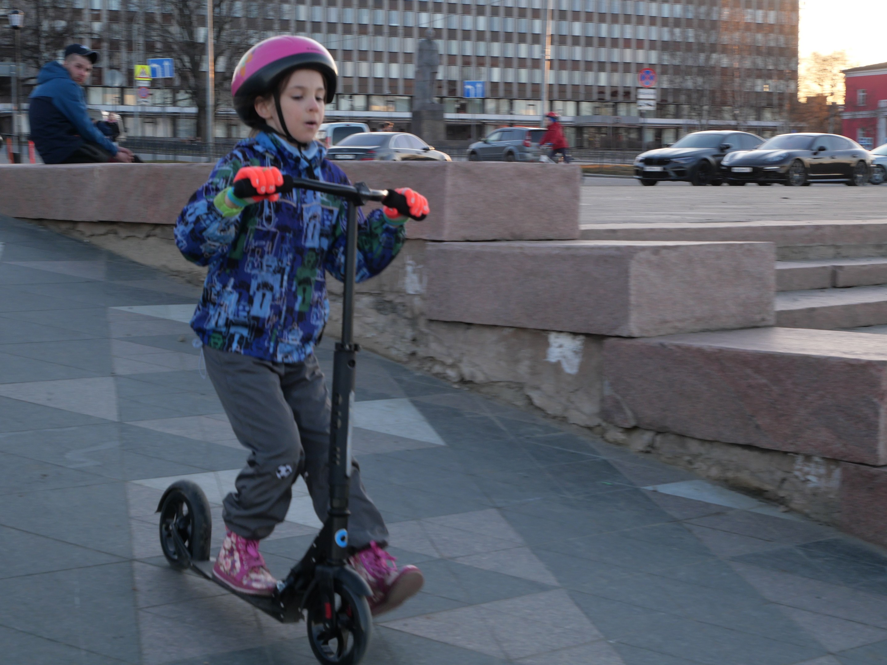 Ein junger Junge in Helm und Handschuhen fährt ein Scooter eine Treppe hinunter auf einem Gehweg mit Fahrzeugen, Menschen, Bäumen, Pfosten, Brettern, Gebäuden und einem klaren blauen Himmel im Hintergrund.