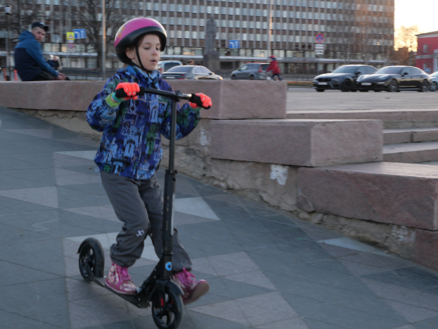 Ein junger Junge in Helm und Handschuhen fährt ein Scooter eine Treppe hinunter auf einem Gehweg mit Fahrzeugen, Menschen, Bäumen, Pfosten, Brettern, Gebäuden und einem klaren blauen Himmel im Hintergrund.