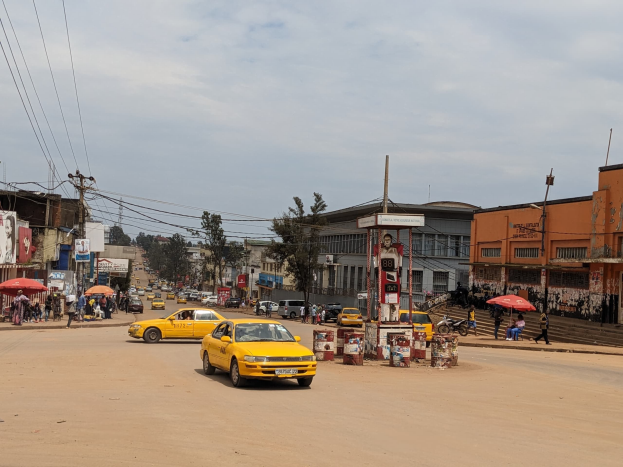 Ein gelbes Taxi fährt eine belebte Stadtstraße mit Gebäuden an den Seiten hinunter, während Fußgänger mit Schirmen auf dem Gehweg unter einem bewölkten Himmel gehen.