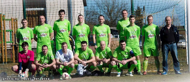 Eine Gruppe von Männern in grünen T-Shirts steht und hockt auf einem Fußballfeld mit einem Ball vor ihnen, einem Torpfosten rechts daneben und einem Hintergrund aus Bäumen, Gebäuden, Pfählen, Fahrzeugen und dem Himmel. Der Text "Bundesliga 1-2-3-4-5-6-7-8-9-10-11-12-13-14-15-16-17-18-19-20-21-22-23-24-25