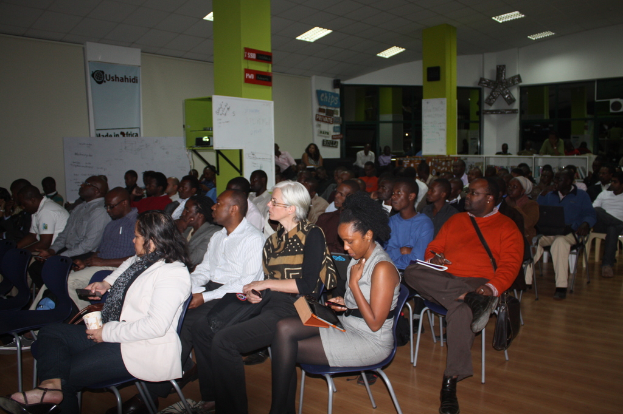 Ein Klassenzimmer mit vielen Menschen auf Stühlen, zwei grüne Säulen mit daran befestigten weißen Tafeln und eine grüne Wand im Hintergrund mit einem Banner und weißen Papieren.
