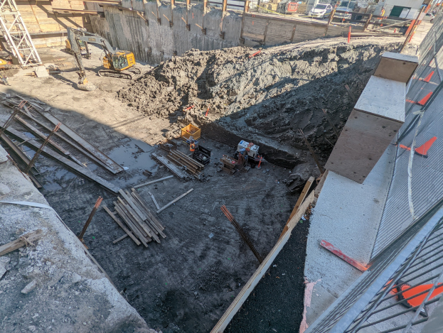 Baustelle mit einem großen Schutthaufen, Kränen, Holzstangen, einem Maschendrahtzaun auf der rechten Seite und Hintergrundelementen wie Gebäuden, Fahrzeugen, Polen, Bäumen und Himmel.