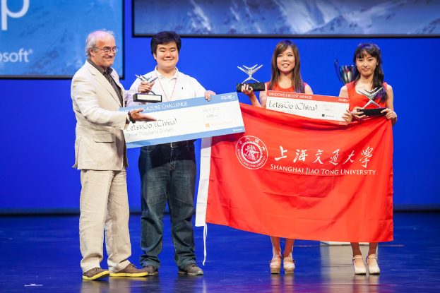 Vier Personen stehen auf einer Bühne, jede hält Auszeichnungen, Schecks und eine Fahne, sie lächeln, mit einem digitalen Bildschirm im Hintergrund, der eine Projektion anzeigt.