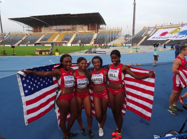 Vier Frauen in Sportbekleidung stehen zusammen auf einer Laufbahn, lächeln und halten amerikanische Flaggen, mit einem Stadion und Himmel im Hintergrund.
