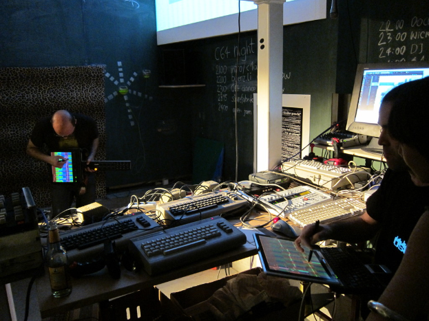 Eine Gruppe von Menschen in Kleidung, die elektronische Geräte halten, mit Tastaturen, System- und Kabelverbindungen, die vor einer Wand mit Text, einem Plakat, einem projizierten Bildschirm und einer Flasche stehen.