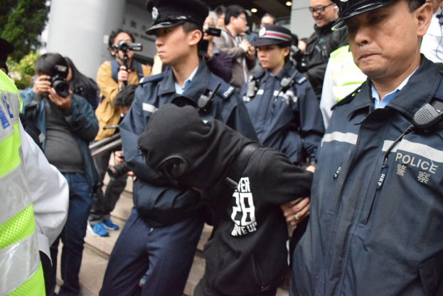 Eine Gruppe von Polizisten steht vor einer Menge von Menschen, einige tragen Mötzen und halten Kameras, mit einem Gebäude mit Säulen und Deckenleuchten im Hintergrund und Bäumen links.