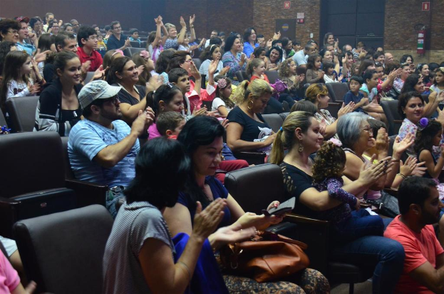 Eine Gruppe von Menschen sitzt auf Stühlen, lächelt, einige halten Gegenstände, tragen Brillen und einer trägt eine Mütze, während einige klatschen, mit einer Wand, Schildern und einem Feuerlöscher oben sichtbar.