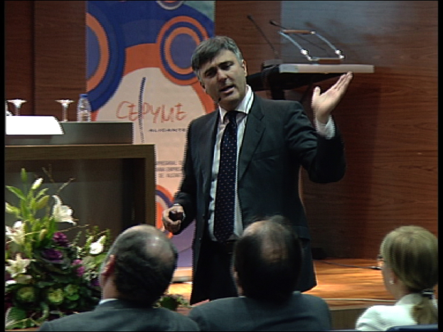 Ein Mann in einem schwarzen Blazer steht an einem Podium mit Mikrofonen, spricht in ein tragbares Mikrofon, mit einer Blumenanordnung und einem Tisch mit einem Buch und einer Wasserflasche in der Nähe, während drei Personen in Stühlen davor sitzen, vor einer braunen Wand und einem bunten Brett mit Text.