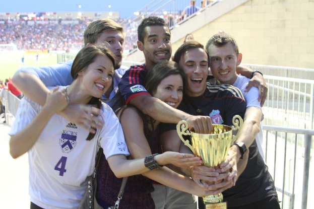 Sechs Menschen, zwei Frauen und vier Männer, die eine Trophäe halten und lächeln, posieren für ein Foto vor einem Eisenzaun, mit einer Wand und einem Zaun oben rechts, Menschen und Lichter im Hintergrund, möglicherweise in einem Stadion.