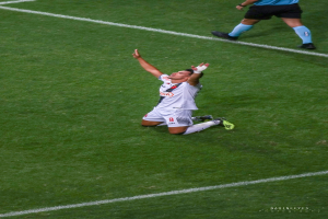 Ein Fußballspieler kniet auf dem Boden und streckt die Arme aus, während er ein Sportoutfit und Schuhe trägt.