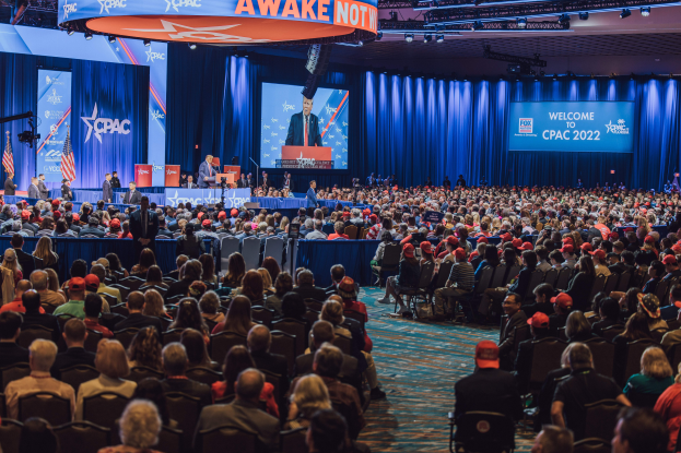 Eine große Menschenmenge sitzt auf Stühlen, die einer Bühne zugewandt sind, mit ein paar Menschen darauf, blauen Vorhängen und einem Bildschirm im Hintergrund und einer orangen Tafel oben, die das CPAC 2020-Event am 25. Oktober anzeigt.