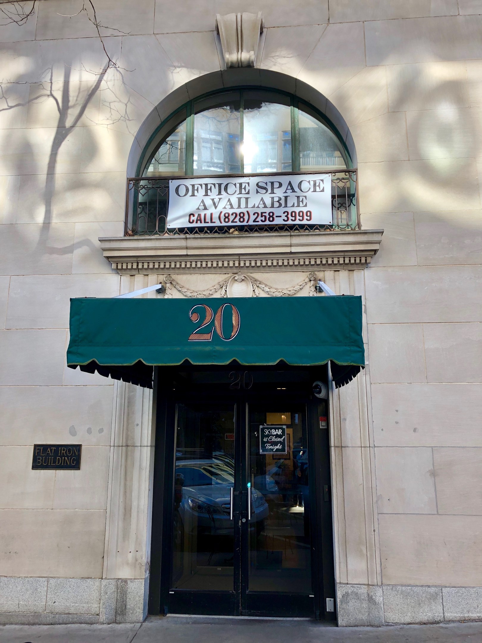 Ein Gebäude mit einer Glastür, einer grünen Markise, einem Schild mit der Aufschrift "Büroräume zu vermieten", einem Geländer und einem Baum auf der linken Seite.