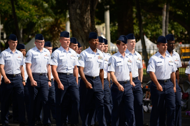 Männer in Uniformen auf einer Straße mit einem Eisenzaun dahinter, Bäume und Pfosten im Hintergrund.