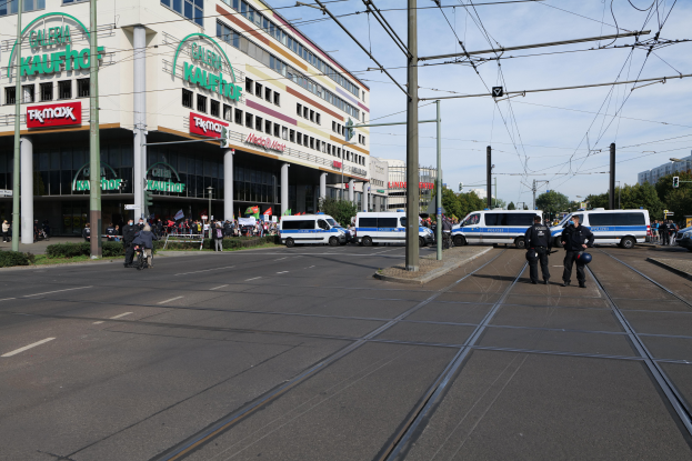 Eine Gruppe von Polizisten steht an der Straßenseite neben einer Tram, mit Fahrzeugen, Strommasten, Gebäuden mit Namenschildern, Bäumen und einem bewölktem Himmel im Hintergrund.
