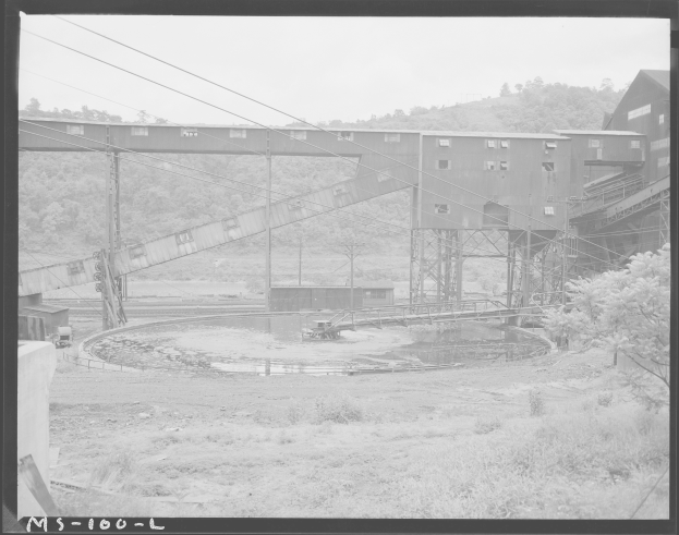 Ein Schwarz-Weiß-Foto eines Kohlebergwerks mit einer Brücke, Pfählen, Drähten, Gebäuden, Bäumen, Pflanzen, Gras und Wasser im Vordergrund und einem Himmel im Hintergrund, mit Text unten.