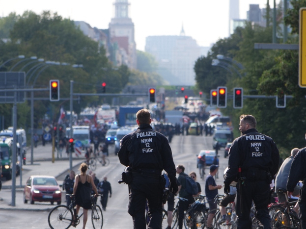 Polizeibeamte mit Fahrrädern auf einer von Bäumen gesäumten Straße mit Gebäuden und einem klaren Himmel im Hintergrund.