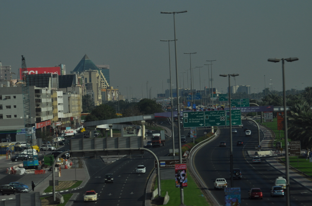 Eine Luftaufnahme einer Stadt mit Gebäuden, Türmen, Kraftfahrzeugen, Werbetafeln, Schildern, Straßen, Bäumen, Straßenlaternen, Menschen, Verkehrszeichen und Informationsschildern unter einem klaren Himmel.
