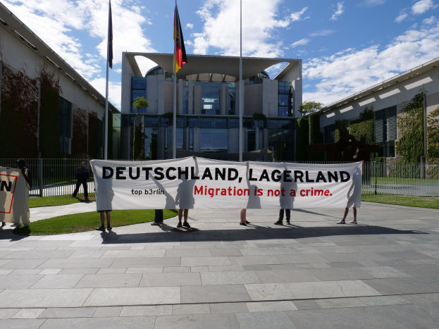 Gruppe von Menschen mit einem Banner "Deutschland, Lagerland Migration ist kein Verbrechen" vor einem Gebäude mit Fenstern, umgeben von Gras, einem Metallzaun, Pflanzen, Bäumen und Fahnenmasten mit Flaggen, unter einem bewölkten Himmel.