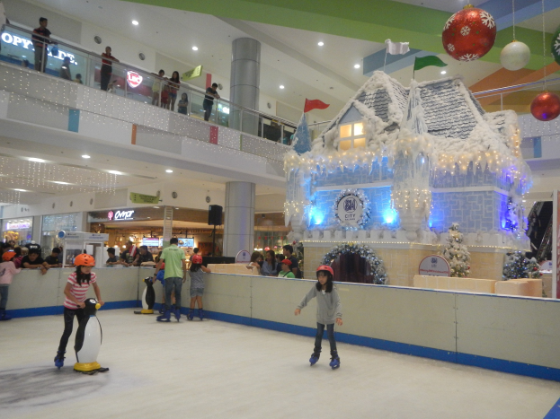 Menschen auf einer Indoor-Eisbahn in einem Einkaufszentrum umgeben von festlichen Dekorationen, darunter ein hausähnliches Gebilde mit Lichtern und eine Menge, die von verschiedenen Ebenen aus zusieht.