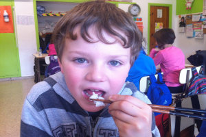 Ein Junge in einer grauen und schwarzen Jacke verwendet Stäbchen, um in einem Klassenzimmer zu essen, wobei andere Kinder hinter ihm sitzen, grüne und weiße Wände, Lehrplakate, eine braune Tür und eine Wanduhr zu sehen sind.