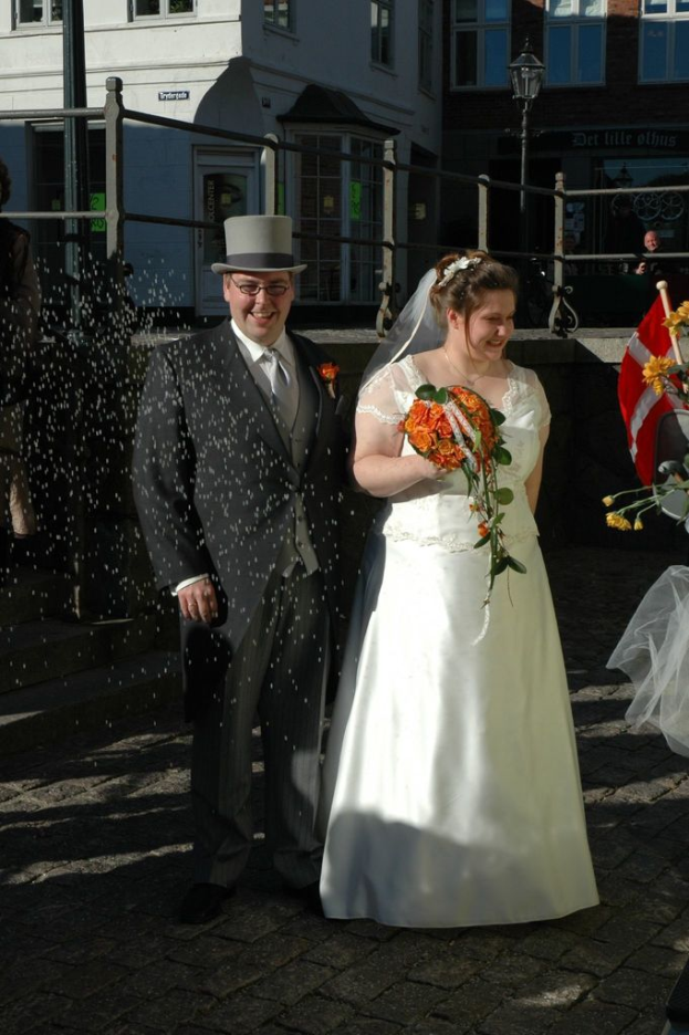 Ein Paar steht zusammen mit der Frau, die einen Strauß hält, einem Zaun und einem Gebäude dahinter und verschiedenen Gegenständen in der rechten Ecke.